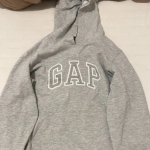 Grå hoodie från GAP - Säljer en klassisk grå hoodie från GAP med stor logga på bröstet. Tröjan har en bekväm passform och en praktisk magficka. Perfekt för en avslappnad stil. Jag är 1,54 lång och tror att det är en barnstorlek
