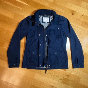 Marinblå fieldjacka Bondelid - Snygg marinblå jacka från Bondelid med dragkedja och knappar. Jackan har flera praktiska fickor och en randig insida. Perfekt för en stilren look. Sitter mer som en S/XS. 
