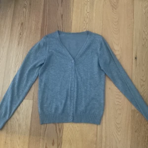 Grå kofta från Emery Rose - Säljer en stilren och bekväm grå kofta från Emery rose i storlek XS. Koftan har en v-ringad design med knappar framtill och är perfekt för både vardag och lite finare tillfällen. Materialet är mjukt och skönt, vilket gör den idealisk för höst och vår