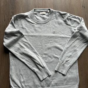 Grå tröja från GANT - en stilren, bekväm grå tröja från GANT. Den är i perfekt skick och har en klassisk design med ribbade muddar vid ärmslut och nederkant. Perfekt för både vardag och lite finare tillfällen. Passar ungdomar i åldern 13-16 år. In köpt i augusti 2024