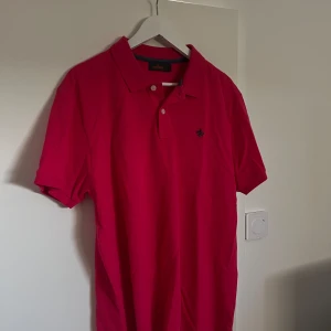 Röd pikétröja från Morris - Säljer en snygg mörkrosa pikétröja från Morris. Den har en klassisk krage och knappar framtill. Perfekt för både vardag och lite finare tillfällen. Tröjan är i bra skick och redo för en ny ägare!