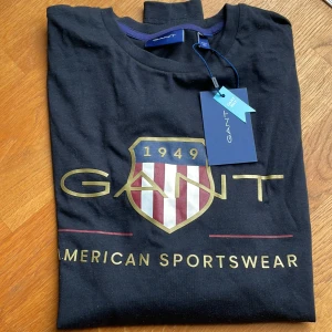 Svart t-shirt från GANT - Säljer en snygg svart t-shirt från GANT med tryck av deras klassiska logga och texten 'American Sportswear'. T-shirten är i nyskick och har en normal passform. Perfekt för en casual look!avser tonåring stl S