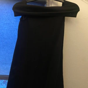 Svart Diana klänning - Snygg svart klänning med offshoulder-design. Perfekt för festliga tillfällen. Klänningen har en elegant och tidlös stil.