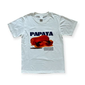 Papaya Tee - Vit t-shirt med tryck av en papaya i fint skick, helt ny och oanvänd.  Trycket är blått och rött och innehåller texten "PAPAYA" och annan lite mindre text.   På baksidan av t-shirten finns inget tryck.
