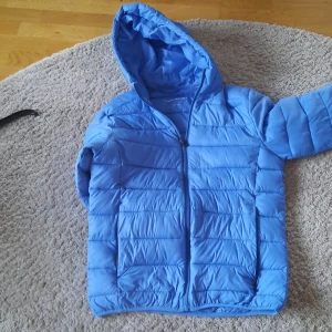 Blå dunjacka med huva - Säljer en snygg blå dunjacka från Lindebergh. Den är perfekt för kyliga dagar med sin pufferstil och har en praktisk huva. Jackan har en dragkedja framtill och är långärmad. Den är i bra skick och redo för nya äventyr! En liten fläck på framsida.