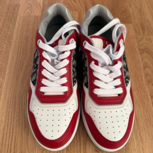 Röd och vit Dior sneakers - Snygga Dior sneakers i rött och vitt med grå detaljer. Skorna har ett coolt mönster på sidan och klassisk snörning. Perfekta för att ge din outfit en lyxig touch. De är i nyskick och redo att bäras till både vardag och fest.