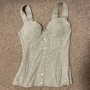 Grå korsettopp från Lindex - Snygg grå korsettopp från Lindex i nyskick. Den har knappar framtill och breda axelband för extra stöd. Baksidan har en smockdetalj som ger en bekväm passform. Perfekt för både vardag och fest! Storlek 75A