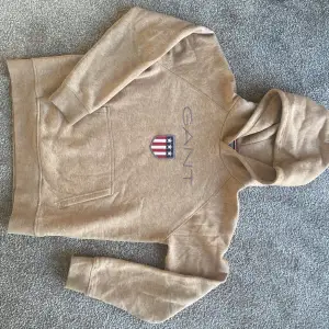 Säljer en snygg beige hoodie från GANT. Den har en klassisk design med GANT-loggan och ett coolt emblem på framsidan. Perfekt för en avslappnad stil och passar bra till både jeans och joggers. Superbekväm och stilren!