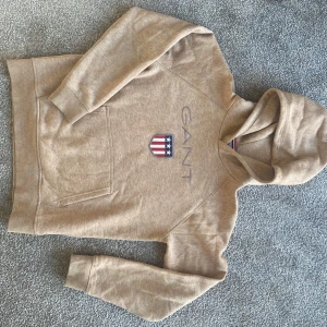 Beige hoodie från GANT - Säljer en snygg beige hoodie från GANT. Den har en klassisk design med GANT-loggan och ett coolt emblem på framsidan. Perfekt för en avslappnad stil och passar bra till både jeans och joggers. Superbekväm och stilren!