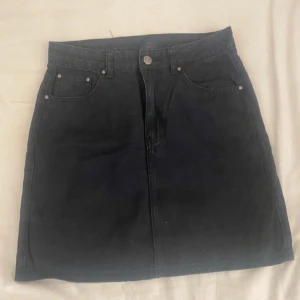 Svart jeanskjol - Snygg svart jeanskjol i klassisk stil. Kjolen har en knapp och dragkedja framtill samt praktiska fickor både fram och bak. Perfekt för både vardag och festliga tillfällen. Passar bra till en t-shirt eller en stickad tröja för en mer avslappnad look.