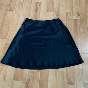 Svart satin kjol från Chiquelle - Svart satin kjol från Chiquelle i en klassisk A-linjeform. Perfekt för en stilren look med sin glansiga yta och mjuka fall. Passar  både till vardag och festliga tillfällen! Aldrig använd 