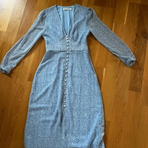 Blå klänning från Adoore - Blå klänning från Adoore med ett  mönster och v-ringad design. Klänningen har långa ärmar och knappar framtill som ger en stilren look. Perfekt för en chic och sofistikerad stil.