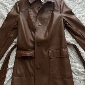 Brun skinnjacka/kappa från Paul Mark - Snygg brun skinnjacka/kappa från Paul Mark med knappar och bälte i midjan. Jackan har en leopardmönstrad insida som ger en cool kontrast. Helt ny med prislapp