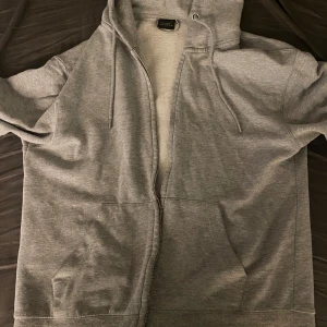 Grå hoodie från Pull&Bear - Säljer en grå hoodie från Pull&Bear med dragkedja och justerbar huva. Perfekt för en avslappnad stil. Den har praktiska fickor framtill och är tillverkad i ett mjukt material.