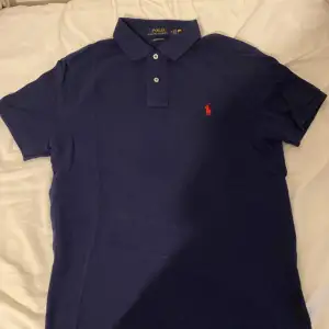 Snygg mörkblå pikétröja från Ralph Lauren med klassisk krage och två knappar. Den har en röd broderad logga på bröstet och är i en slim fit passform. Perfekt för en stilren look.