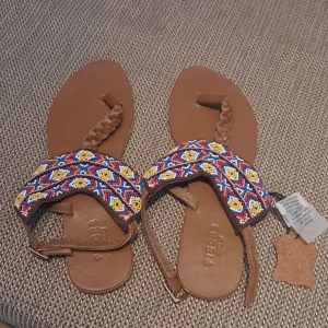 Färgglada sandaler från Pieces - Snygga sandaler från Pieces med färgglatt pärlbroderi i blått, gult och rött. Sandalerna har en flätad rem och justerbart spänne för perfekt passform. Perfekta för sommarens äventyr!