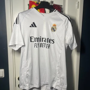 Real madrid fotbollströja - Säljer en vit Real Madrid fotbollströja från Adidas med korta ärmar. Tröjan har klubbens emblem på bröstet och Adidas-loggan i svart. Tillverkad med AEROREADY-teknologi för optimal komfort.