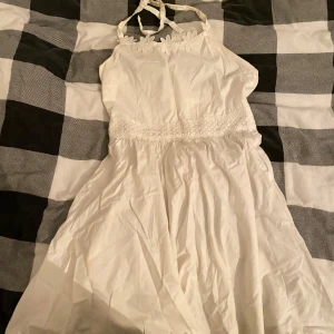 Vit klänning med blommig spetsdetalj - Charmig vit klänning med blommig spetsdetalj vid halsen och midjan. Axelbandslös design med knytband och dragkedja i ryggen. Perfekt för somriga tillfällen.