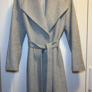 Grå kappa med bälte - Elegant grå kappa med breda slag och knytbälte i midjan. Perfekt för kyliga dagar med sin stilrena design och långa ärmar.
