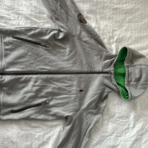 Grå hoodie från Polo Ralph Lauren - Snygg grå hoodie från Polo Ralph Lauren med grönt foder i huvan. Jackan har dragkedja framtill och två fickor med dragkedjor. Märkets ikoniska logga är broderad på bröstet och en patch på ärmen.