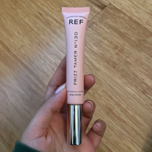Frizz Tamer N°130 från REF - Frizz Tamer N°130 från REF är en hårprodukt i en rosa tub med silverlock. Den innehåller 30 ml och är designad för att tämja frissigt hår. Produkten innehåller quinoa-protein och kokosolja för att ge glans och kontroll.