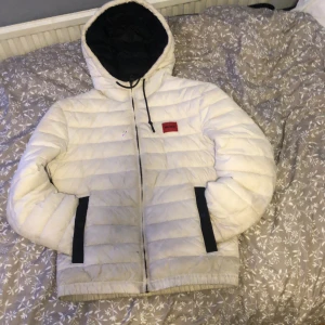 Vit dunjacka från Hugo Vit - Säljer en stilren vit dunjacka från Hugo med svart foder och huva. Jackan har en dragkedja framtill och en röd logotyp på bröstet. Perfekt för kyliga dagar.