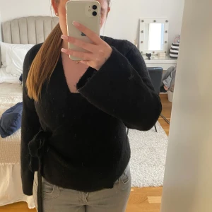 Svart omlottopp med knytband - Snygg svart omlottopp med knytband i midjan. Tröjan har en v-ringad design och långa ärmar. Använd 2-4 gånger och har inga defekter.