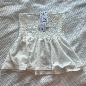 Vit axelbandslös topp - Säljer en söt vit axelbandslös topp med smockdetaljer och volang nedtill. Aldrig använd!! Perfekt för sommardagar och kan enkelt matchas med jeans eller kjol. Skriv för fler bilder eller om ni har några frågor💗