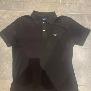 Svart pikétröja från GANT - Snygg svart pikétröja från GANT med klassisk krage och knappar framtill. Perfekt för en stilren look. Diskret logga på bröstet.