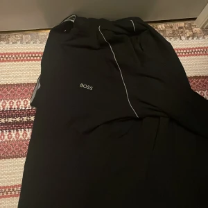 Svart huvtröja från Boss - Snygg svart huvtröja från Boss med dragkedja framtill. Tröjan har en stilren design med vita detaljer och en bekväm grå insida i huvan. Perfekt för en avslappnad stil.