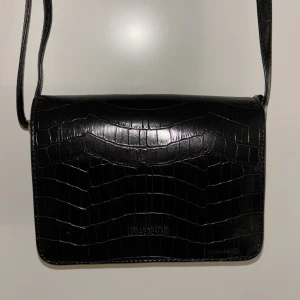 Svart axelväska med krokodilmönster - Snygg svart axelväska med krokodilmönster och justerbar axelrem. Väskan har en praktisk dragkedja på baksidan för extra förvaring. Perfekt för att ge en stilren touch till din outfit.