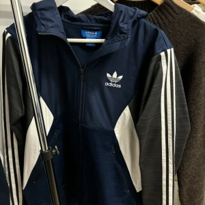 Adidas vindjacka/kofta - Säljer en blå vindjacka/kofta från Adidas med vita detaljer och klassiska ränder på ärmarna. Jackan har dragkedja och en hög krage för extra skydd mot vind. Utöver det har den en inbyggd bandanda (se bild 3) vilket är sällsynt. Perfekt för sportiga aktiviteter eller en casual look. Nypris: 799kr