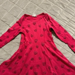 Rosa klänning med hjärtan - Söt rosa klänning med glittriga hjärtan över hela tyget. Klänningen har långa ärmar och en rund halsringning, perfekt för en lekfull stil.