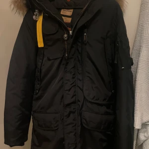 Svart parkajacka från Parajumpers - Säljer en svart parkajacka från Parajumpers med pälsdetaljer på huvan och en gul rem. Jackan har flera fickor och en broderad patch på ärmen. Perfekt för kalla vinterdagar.