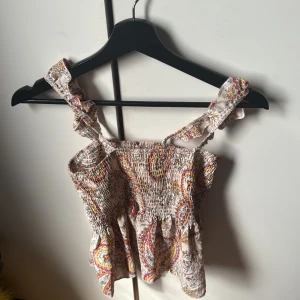 Mönstrad topp med volangdetaljer - Säljer en söt topp med paisleymönster i olika färger. Toppen har volangdetaljer vid axelbanden och en smockad design som ger en bekväm passform. Perfekt för sommarens varma dagar! Från Shein 