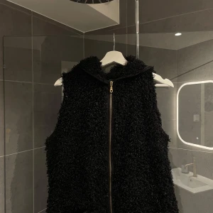 Svart teddy väst  - Snygg svart teddy väst med dragkedja och huva. Perfekt för att hålla sig varm och stilren under kyliga dagar. Västen har en mjuk och fluffig textur som ger en lyxig känsla.