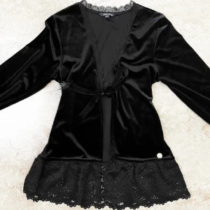 Svart sammetstopp med spetsdetaljer - Elegant svart topp/cardigan i mjuk sammet med långa ärmar och spetsdetaljer vid fållen. Toppen har en knytning framtill och en vacker spetskant längs halsringningen. Använd 1 gång, fint skick men är lite spretad på ena snöret (se bild 4)