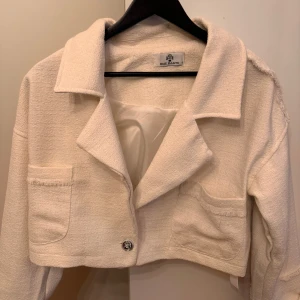 Beige croppad jacka. Med eleganta deltajed!  - Säljer en stilren beige croppad jacka. Jackan har en klassisk krage och två framfickor med dekorativa knappar. Perfekt för en trendig look. Jackan har aldrig använts och är helt ny. 