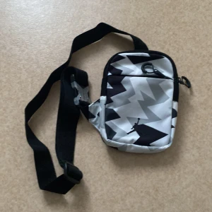 En snygg Jordan väska  - Snygg axelväska med svart och grått mönster och en justerbar rem. Väskan har en dragkedja och ett coolt tryck på framsidan. Perfekt för att bära med sig det nödvändigaste.