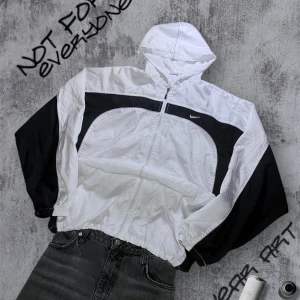 Vit och svart vindjacka från Nike - Snygg vit och svart vindjacka från Nike med dragkedja och huva. Jackan har en sportig look med kontrasterande svarta paneler och en justerbar dragsko i nederkanten. Perfekt för en aktiv livsstil. Lappen är bortklippt men skulle säga L/XL men finns så man kan dra åt längst ner 