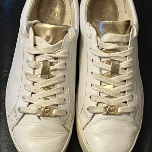 Vita sneakers med gulddetaljer - Snygga vita sneakers från Michael Kors med gulddetaljer på snörningen och tungan. Skorna har en klassisk design med snörning och är tillverkade i skinn med en gummisula.