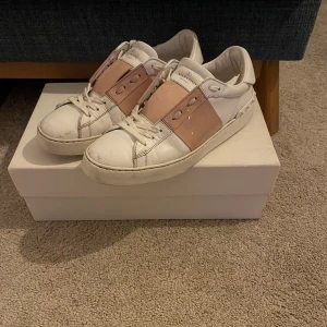 Valentino Garavani sneakers - Lägg bud!  Snygga Valentino Garavani sneakers i vitt med rosa detaljer. Storlek 40 men passar mindre. Skorna har en klassisk design med snörning och en bred rosa rem över ovansidan skorna är i jätte fint skick knappt använda har haft skorna i 2 år men bara använt dom på sommaren och väldigt få gånger då. Perfekta för en stilren look. Nypris 7699kr