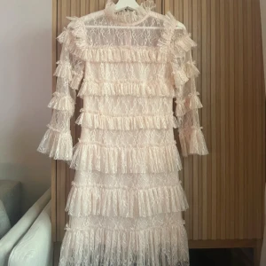 By Malina klänning  - Elegant beige klänning i spets med volanger över hela plagget. Den har långa ärmar och en hög halsringning, vilket ger en romantisk och sofistikerad look. Perfekt för speciella tillfällen. Rosa / Beige färg 