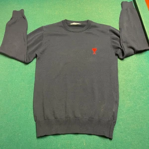 Svart tröja från Ami - Snygg svart tröja från Ami med ett rött broderat hjärta och bokstaven A på bröstet. Tillverkad i 100% bomull, perfekt för en stilren look. Tröjan har långa ärmar och ribbade kanter.