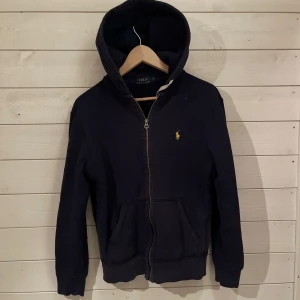 Marinblå hoodie från Ralph Lauren - Snygg marinblå zip hoodie från Ralph Lauren med dragkedja och ett klassiskt med färgen av gul på logga på bröstet. Perfekt för en avslappnad stil till t.ex våren eller hösten, med praktiska fickor framtill och en bekväm huva. Hör gärna av er om ni undrar något om min Polo Ralph Lauren hoodie! Min Polo hoodie är ÄKTA och är i bra skick inget tecken på att den har blivit skadad. Pris ej hugget i sten!