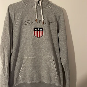 Grå hoodie från GANT - Säljer en snygg grå hoodie från GANT med broderad logga och emblem på framsidan. Tröjan har en känguruficka och justerbar huva med snören. På baksidan finns en röd och vit randig detalj som ger en sportig look.