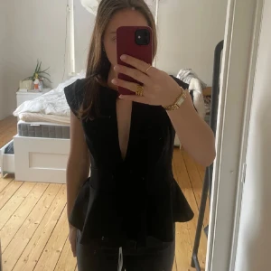 Svart topp med volangdetalj - Snygg svart topp med volangdetalj i midjan och djup v-ringning. Ärmlös design som ger en elegant look. Perfekt för att klä upp en outfit.