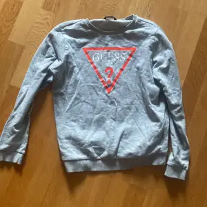 Snygg grå sweatshirt från Guess med det ikoniska röda och vita logotyptrycket på framsidan. Tröjan har långa ärmar och en rund halsringning. Perfekt för en avslappnad stil.