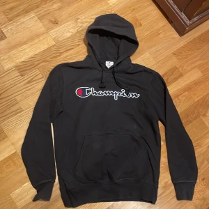 Svart hoodie från Champion - Säljer en svart hoodie från Champion med det klassiska logotyptrycket på bröstet. Tröjan har en justerbar huva och en praktisk magficka. Perfekt för en avslappnad stil.