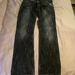 Mörkblå jeans från Lee - Säljer ett par mörkblå jeans från Lee med slitna detaljer på bakfickan. Byxorna har en klassisk femficksdesign och en rak passform. Perfekta för en avslappnad stil.